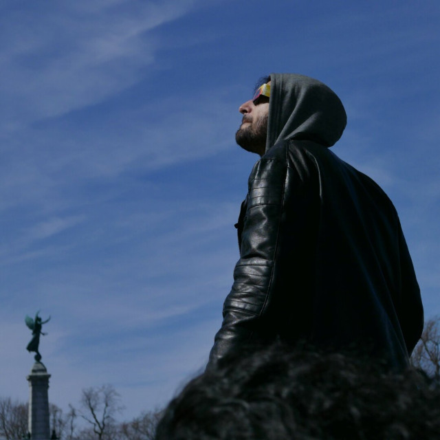 Man wearing hooded leather jacket &amp; eclipse glasses, blue sky &amp; statue in background
