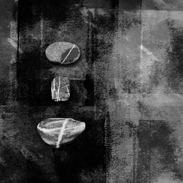 Black and white image of three small water worn stones, all displaying streaks of quartz, on a dark slate, textured background.