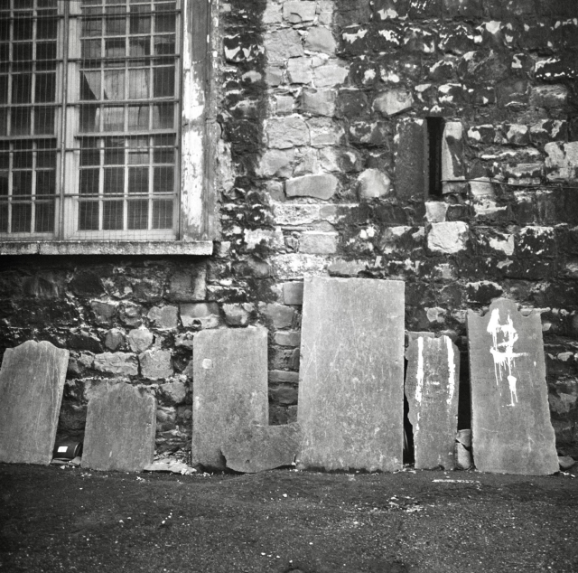 A black and white film photo. A series of very old tombstones are seen leaning against the St. Michan’s stone church wall in Dublin, Ireland. Thursday, October 26, 2023.