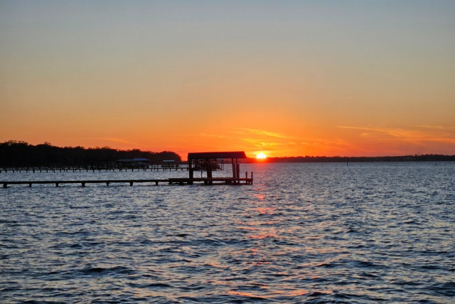 A big bright yellow setting sun has reached the horizon along a vast river, glowing beyond piers and boat docks that line the river's shoreline. Colorful orange shades surround the sun, casting their shades across the horizon and a faint reflection upon the river below.