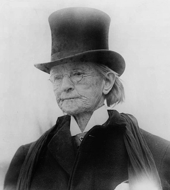 Dr Mary Edwards Walker in later life. She is wearing a wing collar shirt with a cravat, a heavy overcoat and a top hat. She is a white woman with white hair.