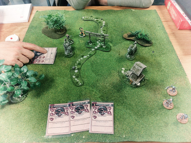 A 60×60 cm field of fake grass, with miniatures and terrain pieces on it. To the right is a hand pointing at a card describing some of the rules of the game.