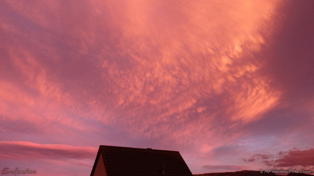 A photo of a cloud system above a rooftop. Beneath the clouds is an odd pattern, with blister-like bulbs lit from the left by the rising sun. This has produced a strong pinkish-orange colour across the formations, making them stand out more. It's quite an unearthly scene.