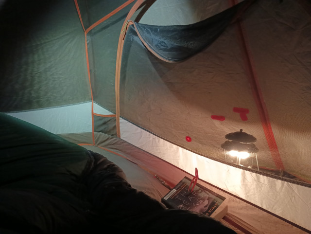 A color photo from inside a small tent. It's night. There's part of a sleeping bag visible in the lower left of the frame. The top of a round tent door is about a quarter open to allow heat from a gas Coleman lantern in the vestibule to enter the tent. There's a book on the floor by the door. It's title is: "The Bears Ears."
