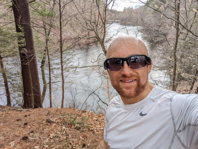 A selfie of me on a forested hill overlooking a wide bend in the Charles River on an overcast day. Forest lines both sides of the Charles as it takes a bend to the right in the distance. I'm a bald, middle-aged white man with a red beard flecked with white wearing black sunglasses and a white running shirt