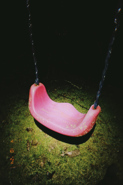 A red swing – of a childrens swingset – sits empty – illuminated by the spotlight of my headlamp– bright green grass behind it