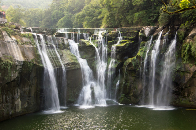A wide waterfall cascading over rocky cliffs into a serene pond, surrounded by lush greenery with spectators on an observation deck to the left.