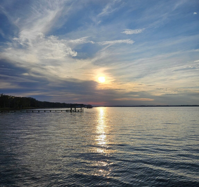 Looking across a wide river where a very hazy, almost blurry sun is approaching the horizon, nearly obstructed by thick bands of cloud cover. With shades of red and blue reflecting on the clouds, and a bright streak of the sun's reflection across the water below.