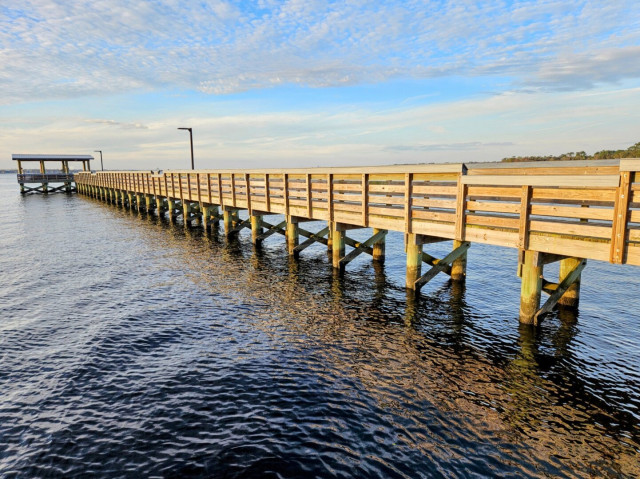 Side view of a long weathered wood fishing pier extending far out into a vast river. The long pier leads to a covered observation &amp; fishing deck overlooking the river. Beneath baby-blue skies lined with rows of cottony fluffy white cloud formations.