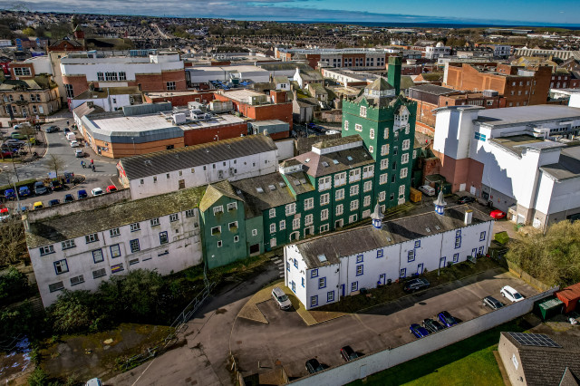 Aerial shot of unusual green and white period six story building with a tower in Workington. Modern building and carparks surround it, with blue cloud speckled sky above