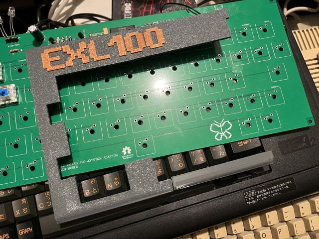 A disassembled computer keyboard with the top panel removed, exposing the green circuit board with key mounts and a few switches. Visible texts include "EXL100," keyboard and joystick adapter information, and "MSX2" branded on the base.