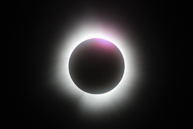 Full solar eclipse photo with a white corona with a splash of pink and red in the upper right in the corona and on the Moon.