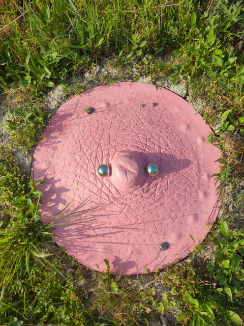 Photograph of a man hole cover, surrounded by grass. The cover has been painted pink like a human nipple, in the middle there's even a shiny metallic piercing through it. The detail on the nipple surface is astounding, right down to the little lines and creases.