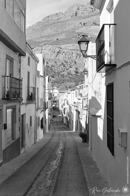 A black-and-white photo looking along the narrow cobbled main street through the old part of Albanchez. White houses are on both sides and a huge mountain is close by, seemingly at the end of the street.