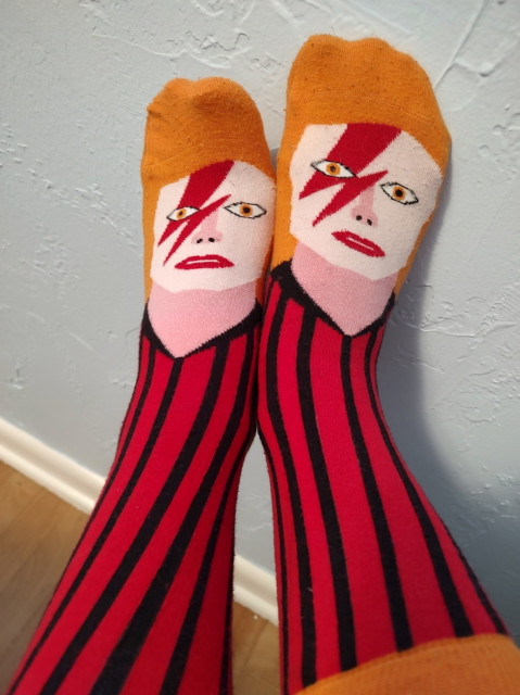 Pic of a woman's outstretched feet in socks knitted to look like David Bowie as Ziggy Stardust. A blue wall and blond hardwood flooring are visible in the background.