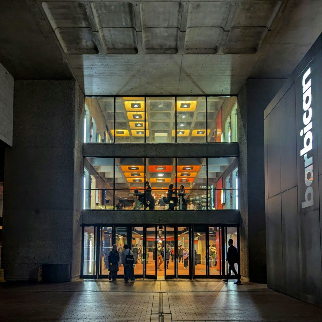 Colour photograph of the main waterside entry to the Barbican Centre in London. There are three floors visible, the ground floor is all glass doors and the two above have floor-to-ceiling windows. The picture is taken at night so the outside is dark, but the inside is fully lit up. On the ground floor figures entering and leaving are silhoutted against the brighter interior, where there are many people moving around. On the floor above, orange light settings can be seen above two tables in the window, at which three people are seated. There is no-one visible on the floor above that, only yellow square sections of the ceiling. All the walls and ceilings visible are concrete, other than where colour has been added around light fixtures. To the right of the entrance is a large black slab with 'barbican' running up the nearest edge, it is lit from behind but it appears as though the bulb behind 'bar' has gone out, because it's noticeably darker than the rest of the word.