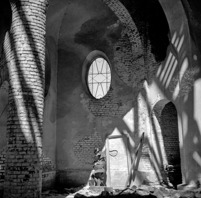 Black and white image of the interior of an abandoned church. The plaster has flown off and bricks are visible. The upper part of the vault is in the form of arches. There is an ellipse-shaped window in the wall.
Sunlight falls on the walls from behind the observer through the dilapidated roof and creates shadows on the walls