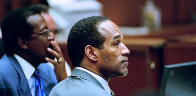 A Black man dressed in a suit sits in a courtroom and is listening to testimony. 
