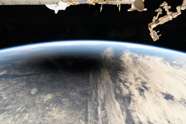 The Earth as seen from space, with parts of the ISS at the top of the picture. A circular shadow caused by the Moon during a total solar eclipse covers the landmass, while clouds are seen to the right of the shadow.