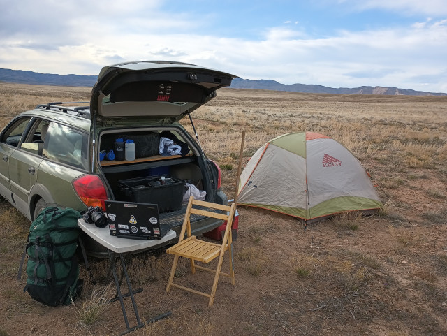 A lone car camp on a relatively flat plain with short dry grasses. On the left is a light green car (Outback) with the rear hatch open. There's a small table on the left with a laptop and camera. A wood chair is behind the table. And there's a small grey a light green tent on the right side of the frame.