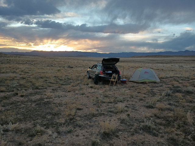 A sunset behind some very distant mountains that are low on the horizon. To the right of the sunset is a car with its rear hatch open. To the right of the car is a small tent. The car and tent are alone on a wide vast light brown plain.