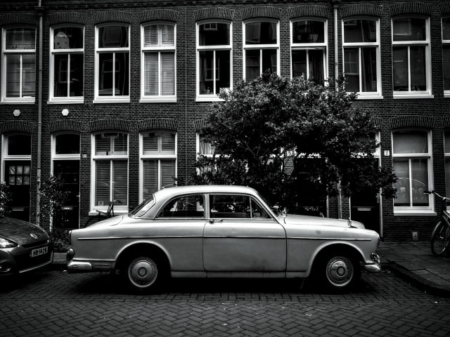 Black and white photo taken across the street from a 1960s Volvo automobile parked in front of a brick block of flats.