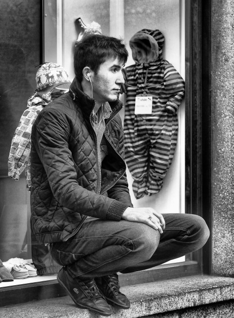 Ein schwarz-weißes Hochkant-Foto.
Ein junger Mann sitzt auf Zehenspitzen, in der Hocke, auf einer Fensterbank eines Schaufensters. Er schaut verträumt geradeaus und hat kabelgebundene in-Ear-Kopfhörer in den Ohren und hört sehr wahrscheinlich Musik.