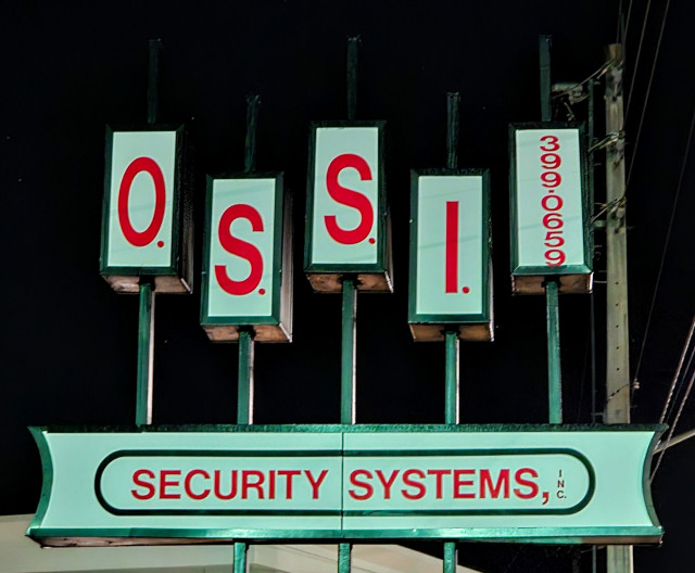 Late night looking up at signage for "OSSI Security Systems, Inc" using a pattern reminiscent of the 1960's era advertising style. With small white rectangles, each containing one letter and appearing at different heights as if they had slid out of place on their pole. With the company name and initials in red letters against the white sign background and reflecting a tinge of green from a nearby traffic light.