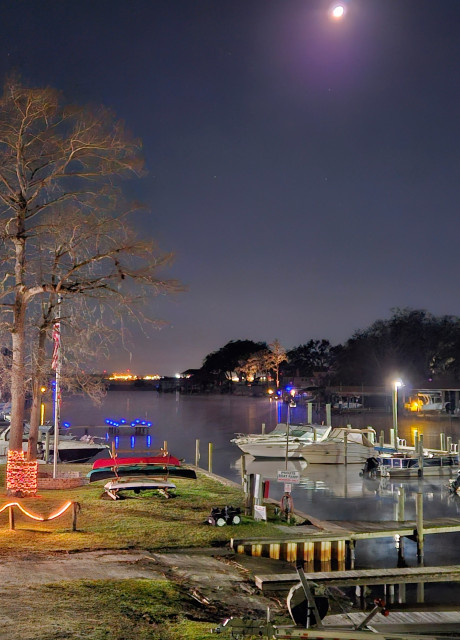 Late night overlooking a small marina at the mouth of Goodby's Creek and the Saint Johns River beneath a glowing partial moon. Red decorative lights outline portions of the marina's shoreline features. Blue lights outline a nearby dock and a smaller one on the far shoreline across the water. Yellow lights appear at the rear of some boats on both sides of the water. Bright city lights can be seen far off in the distance beyond the creek, and the vast river the creek flows into. All brightly illuminated and reflecting upon the water below a blue night sky.