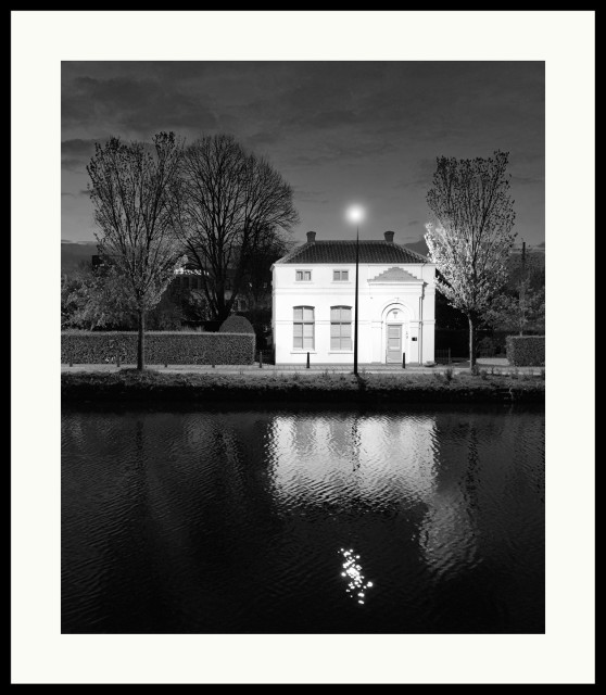 b&w - single house on the waterfront. a blurred reflection is seen in the water. scene is primarily lit by one street light. shot during blue hour.