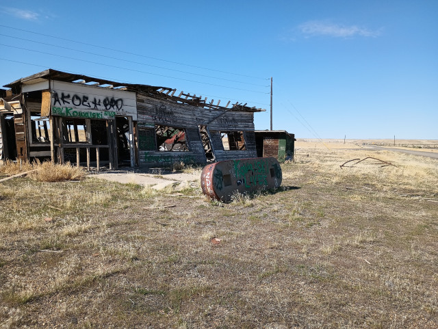 Color photo of an abandoned gas station / cafe. It is in very poor shape and will likely completely collapse in the next couple of years. The gas pump island has been turned on it's side and someone painted "Kowalski Lives" on it. There's short dry grass surrounding the building and an empty road stretches off into the distance. 