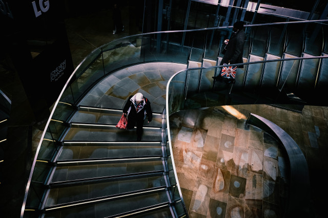 A colour photo with a high-contrast cinematic feel looking down a concrete and steel staircase at the One New Change shopping centre in London on two people descending the stairs, one a silver-haired older man carrying a red and white shopping bag, the other a black guy wearing a black woollen hat, white wired earbuds, and an M&S shopping bag with the union flag on the side.