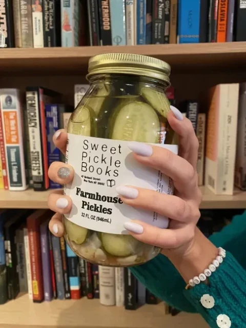 person with pretty nails holding a jar of farmhouse pickles from sweet pickle books in front of a full bookshelf.