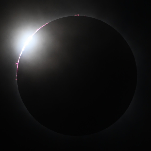 Diamond-ring effect seen through thin clouds, with red chromosphere and prominences visible on the upper left part of the Sun and a bright spot at upper left just before the start of totality.