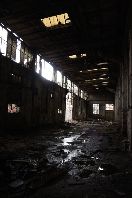 Interior of very dark, narrow building - ground covered in mud, with puddles reflecting the glassless windows and doorless entryway to reveal a glimpse of derelict industrial buildings beyond. Yellowed skylights above add to the general sense of abandonment.