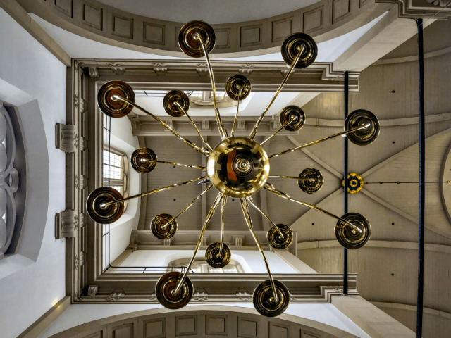 Looking up to the ceiling of a large church. The photo is taken directly under an elaborate brass(?) lamp with a large sphere is the middle.
