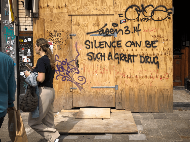 A large sheet of plywood covering a storefront with the graffiti text "Laser 3.14 SILENCE CAN BE SUCH A GREAT DRUG".
On the left of the frame a young woman in beige pants can be seen walking away.