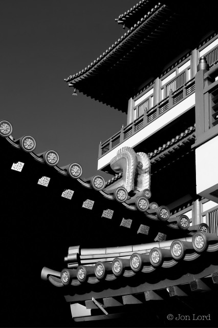 A High Contrast Black And White Photo In Portrait Format. The Image Shows An Oblique View Of The Front Facade Of The Buddha Tooth Relic Temple. The Bottom To Half Way Up The Photo Shows The Eves Of The Sloping Roof. We Can See The Ends Of The Beams Supporting The Glazed Tiles, Only The Edges Of The Tiles Are In View, They Are Embellished With A Round Sun Burst Where The Tiles Join. The Upper Half On The Right Side Is The Facade Built In The Chinese Buddhist Architectural Style Of The Tang Dynasty. There Are More Examples Of Ornately Finished Sloping Roofs, Balconies, And Shuttered Windows. On The Upper Left Side Is A Jet Black Sky With No Clouds Present. 

Singapore, 2015


