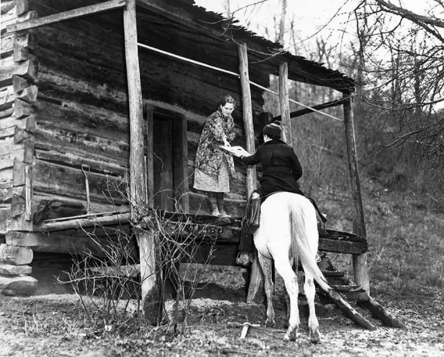 Black and white photo of a pack horse librarian delivering books to a woman standing on the high front porch of a cabin in rural Kentucky in 1938.