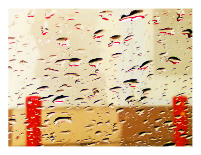 rainy morning, out of focus through my car windshield. two red parking bollards stand at opposite ends of the picture in front of a building wall that's painted and patched in several shades of brown and beige. in-focus raindrops reflect red, white and brown colors.