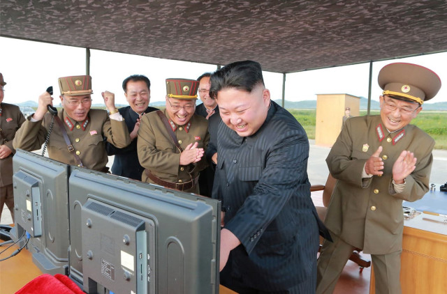 Kim Jong-Un standing behind two LCD monitors, pointing at one of the screens and smiling. Around him various military officers and functionaries are cheering and clapping him.