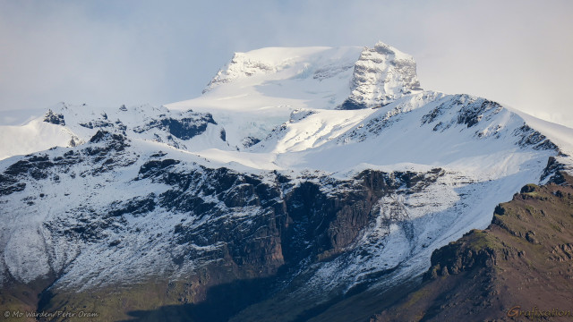 A photo of a mountain peak, lit from the right and under a cloudy sky. The peak is almost completely covered in snow and ice apart from the faces which have been warmed by sunlight or stripped by the wind. Where the rock surface is visible, it's a dark brown colour. At the top is a curved wall, similar to the vent created by a volcano but only about fifty percent of it remains. This oddly shaped half ring is filled with snow but no glacier is apparent. It's beautiful, but in an intimidating way.