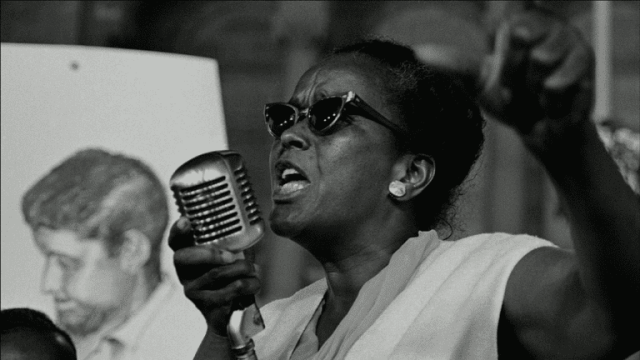 Photo of Ella Baker speaking at a microphone. She is a black woman wearing sunglasses.