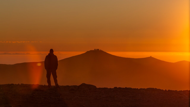 A photograph showing the silhouette of a person standing on rocky terrain in the foreground and a mountain range in the background. Two peaks are visible, one in the centre of the image and another one farther to the right, and on each of them are small constructions. The photo was taken during the sunset so the whole image is washed in shades of orange, making it difficult to distinguish details.