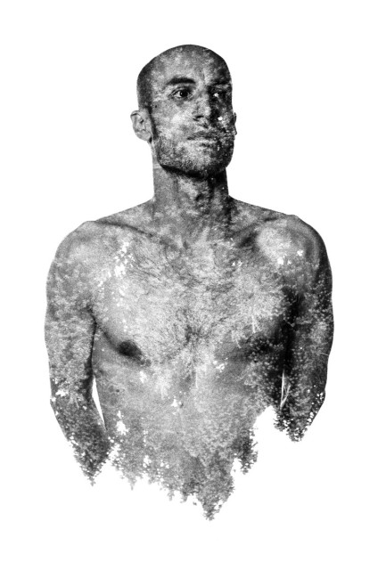 A bald man standing naked with his arms behind his back. His skin is mottled with tree leaves and the lower part of his torso fades into leaves. Black and white. 
