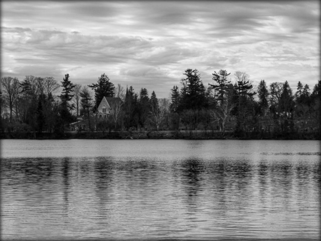 Looking across a small lake towards the other side. Large trees and a small building can be seen. Clouds fill the sky in this moody black and white image.