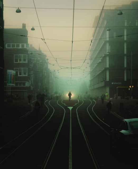 Photography. A color photo of a morning street scene in foggy Amsterdam. In the center is a cyclist who can be seen in the distance 
behind long, parallel streetcar tracks. The cyclist is illuminated from behind by yellow light. To the left and right, rows of houses are bathed in green/grey. Above them hang the power cables of the streetcar. The photo lives from the seemingly endless rails in the foreground and its greenish blurred color. It radiates tranquillity before the coming hustle and bustle of the city.
Info: The photographer comments:
"As I picked up my camera to take the shot, the truck drove over a bump and its headlights illuminated the cyclist for a split second, and at that exact moment I pressed the shutter."