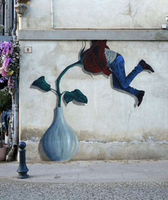 Streetartwall. A funny mural of a man hanging in a huge flower has been painted/glued to the outside wall of a flower store. A gray-green flower vase with a large dark red rose becomes the man's undoing. He hangs with his head up to his shoulders in the flower and wriggles his legs in the air. The man is wearing jeans, a striped T-shirt and black shoes. (The photo shows purple flowers in front of the store on the left).