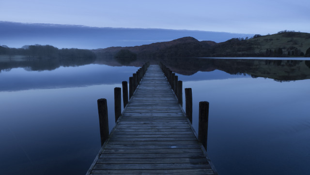 A long wooden jetty runs away from the camera into a calm mirror like lake. A bank of cloud sit on the horizon and mist nags on the banks of the lake. The picture has a blue tint from the time of day.  