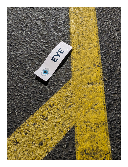 night by streetlight. overhead view of black asphalt with yellow painted lines. an oblong piece of white, crumpled, coated paper with an illustration of a blue eye and the the word EYE lies at an angle and casts a small shadow.